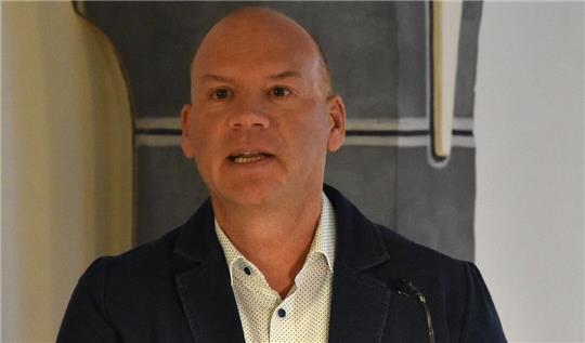 Mann mit grauem Haar trägt ein weißes Hemd mit Muster und einen dunklen Blazer vor einer Wand mit schwarzem und weißem Muster