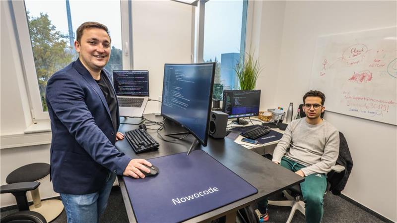 Ein Büro mit mehreren Schreibtischen und Monitoren. Links steht ein Mann an seinem Schreibtisch, rechts sitzt ein anderer auf seinem Schreibtischstuhl.