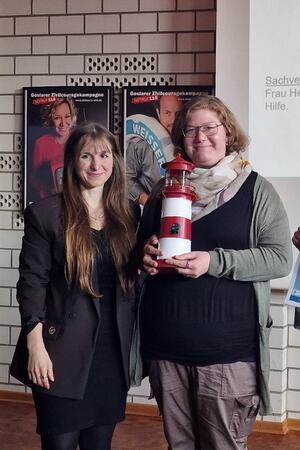 Zwei Frauen stehen nebeneinander, eine hält ein rot-weiß gestreiftes Modell eines Leuchtturms.