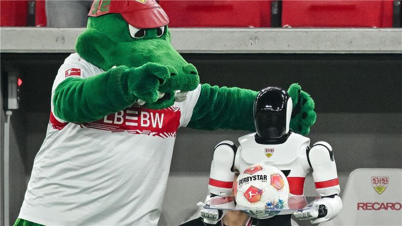 Maskottchen Fritzle neckt einen Roboter mit dem Spielball am Spielfeldrand.