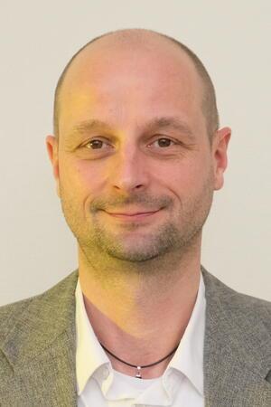 Porträtaufnahme eines Mannes mit Glatze, der ein weißes Hemd und ein graues Sakko trägt sowie eine Halskette mit kleinem Anhänger.