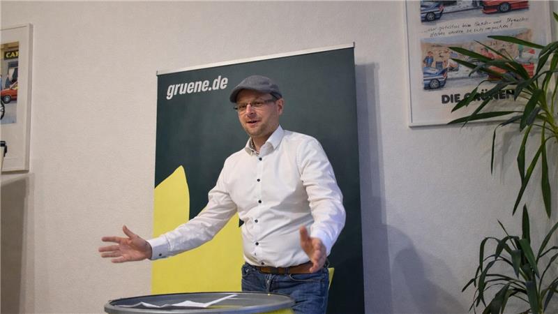 Mann mit weißem Hemd und grauer Mütze gestikuliert vor grün-gelbem Banner mit Schriftzug 'gruene.de'.