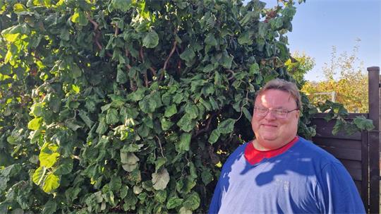 Mann mit Brille und blauem Pullover steht vor einer hohen, dicht bewachsenen grünen Hecke im Freien