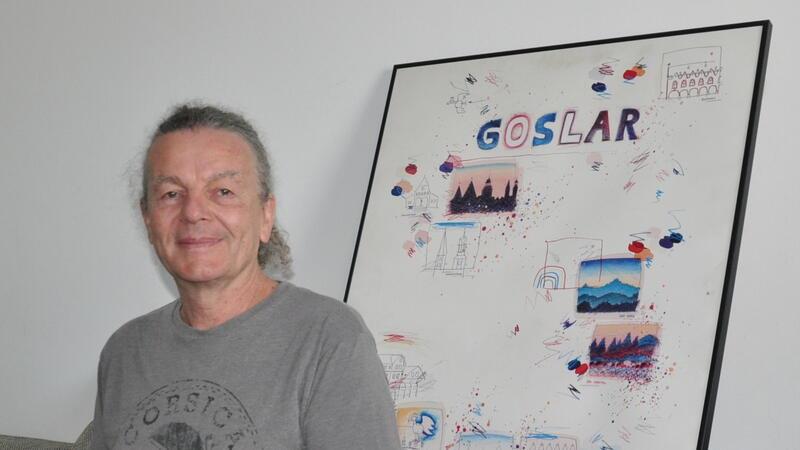 Ein Mann mit grauem T-Shirt sitzt vor einer Wand, an der ein großes, gerahmtes Bild mit dem Schriftzug "Goslar" lehnt.