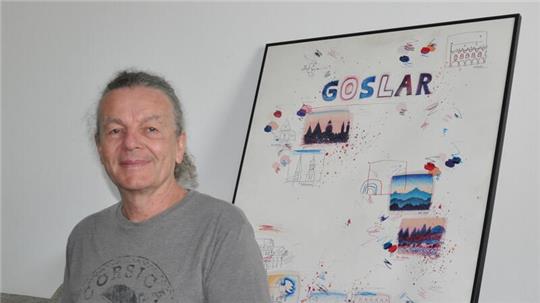 Ein Mann mit grauem T-Shirt sitzt vor einer Wand, an der ein großes, gerahmtes Bild mit dem Schriftzug "Goslar" lehnt.