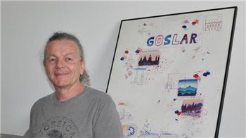 Ein Mann mit grauem T-Shirt sitzt vor einer Wand, an der ein großes, gerahmtes Bild mit dem Schriftzug "Goslar" lehnt.