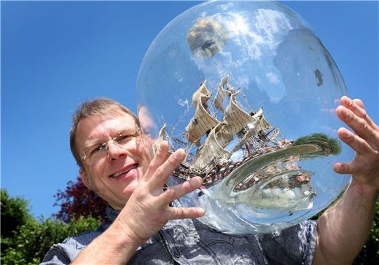 Matthias Schultz hält ein von ihm gebautes 40 Zentimeter hohes und 38 Zentimeter langes Buddelschiff welches in einem 50 Liter fassenden Gärballon hängt.