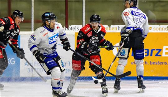 Zwei Eishockeyspieler in weißen Trikots und zwei Spieler in schwarzen Trikots versuchen den Puck zu kontrollieren.