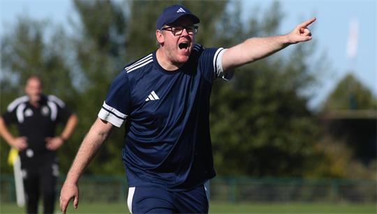Mann in blauem Sportoutfit und Mütze schreit und zeigt mit ausgestrecktem Arm nach rechts auf einem Sportfeld