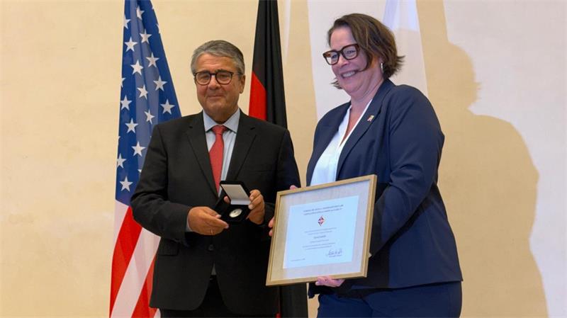 Zwei Personen in formeller Kleidung stehen vor den Flaggen der USA und Deutschlands, eine Person hält eine Urkunde, die andere eine Medaille