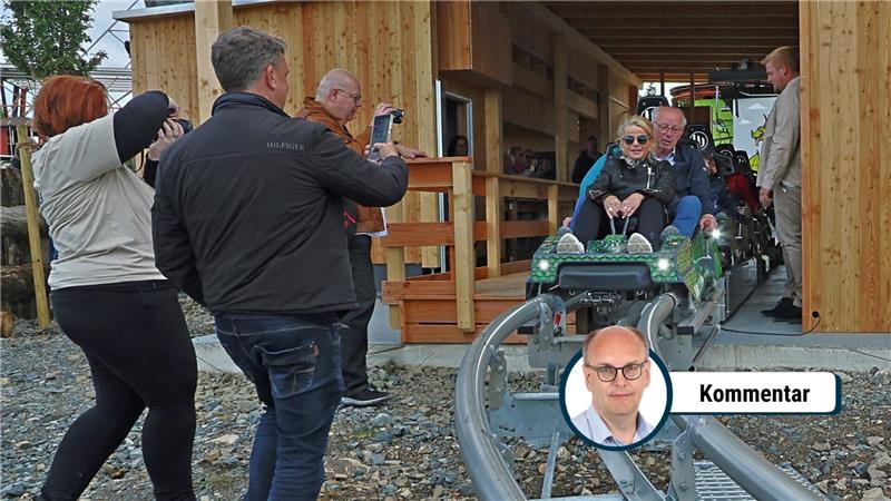 Mehrere Personen beobachten und fotografieren zwei Personen in einem kleinen Wagen auf einer Schienenbahn vor einem Holzgebäude