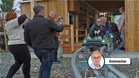 Mehrere Personen beobachten und fotografieren zwei Personen in einem kleinen Wagen auf einer Schienenbahn vor einem Holzgebäude