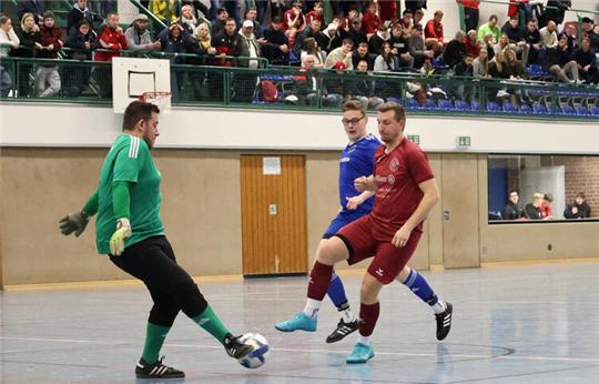 Hallenszene mit mehreren Fußballspielern, ein Spieler in grün hält den Ball, andere Spieler in rot und blau laufen auf ihn zu, Zuschauer auf Tribüne im Hintergrund.