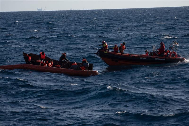Mehrere Migranten sitzen in einem Boot im Mittelmeer während Rettungskräfte versuchen ihnen zu helfen.