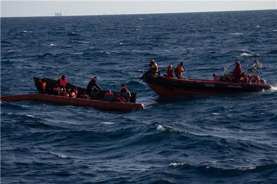 Mehrere Migranten sitzen in einem Boot im Mittelmeer während Rettungskräfte versuchen ihnen zu helfen.