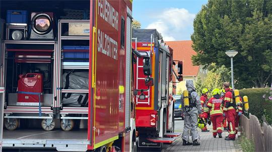 Mehrere Feuerwehrfahrzeuge stehen an einer Straße, Feuerwehrleute in Schutzanzügen und Atemschutzgeräten gehen entlang eines Zauns