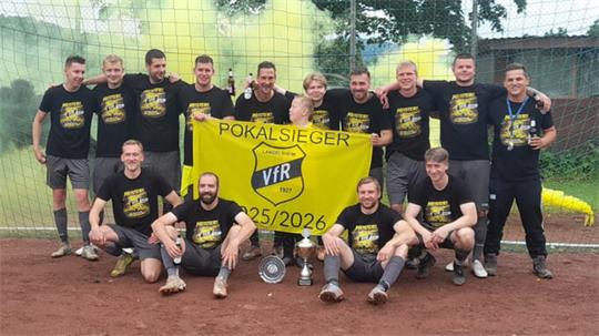 Mannschaft in schwarzen T-Shirts posiert auf rotem Sportplatz vor grünem Netz mit gelbem Banner 'POKALSIEGER VfR 1927 25/2026' und Pokalen