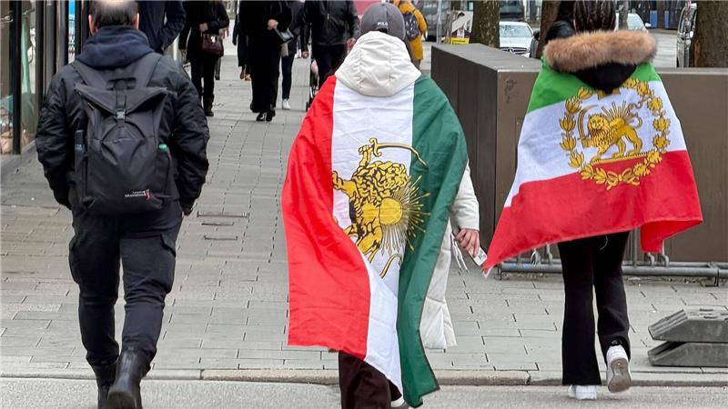 Menschen, die verschiedene ehemalige Flaggen des Iran umhaben, gehen durch die Münchner Innenstadt.