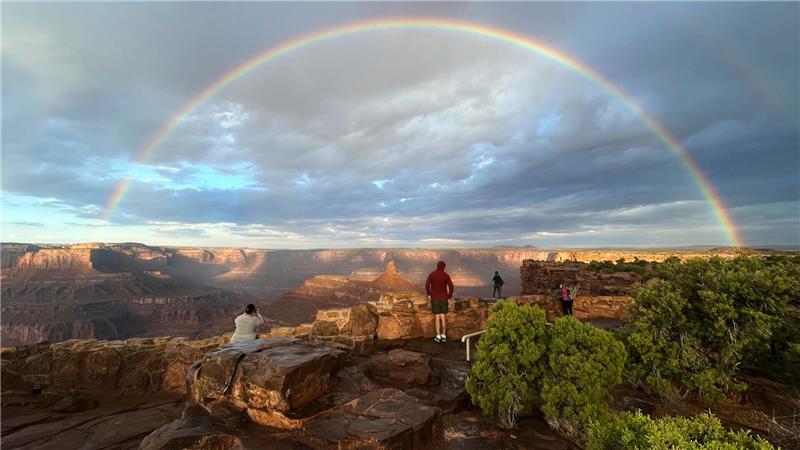 Menschen stehen an einem Aussichtspunkt, während ein doppelter Regenbogen am Himmel über dem Dead Horse Point State Park in der Nähe von Moab in den USA leuchtet.