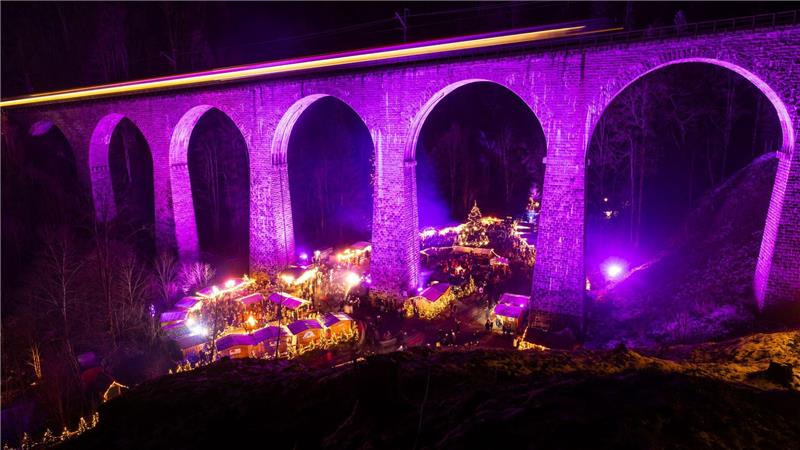 Menschen stehen auf dem Weihnachtsmarkt in der Ravennaschlucht, während über ihnen ein Zug über das Eisenbahn-Viadukt fährt. 