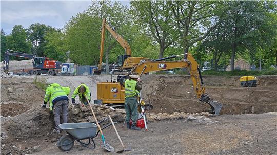 Bauarbeiter in Warnkleidung bedienen Bagger und Schaufeln auf einer Baustelle mit Erde und Bäumen im Hintergrund