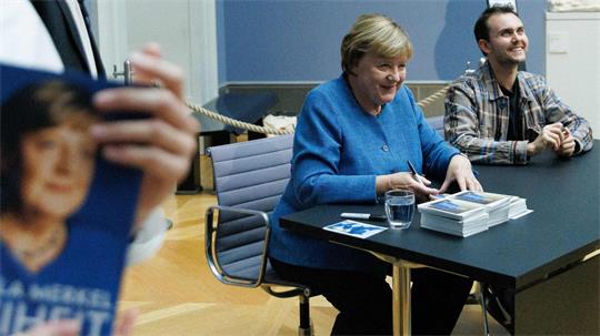 Merkel signiert Kunstpostkarten im Bode-Museum neben dem Maler ihres dort ausgestellten Porträts, Jérémie Queyras. 