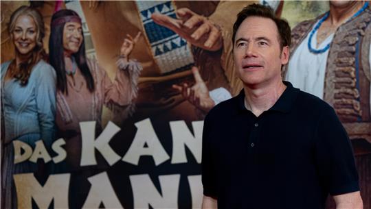 Ein Mann steht vor einer Wand mit einem großen Plakat, auf dem der Text 'DAS KAN MAN 'und viele Personen zu sehen sind.