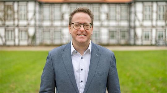 Mann mit Brille und blauem Sakko steht lächelnd vor einem historischen Fachwerkhaus