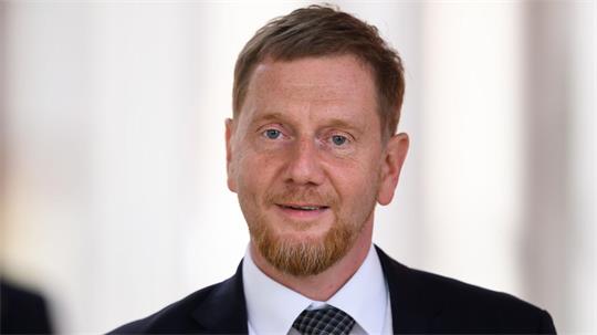 Porträt eines lächelnden Mannes mit kurzem roten Haar und Bart, der einen dunklen Anzug und ein kariertes Hemd trägt
