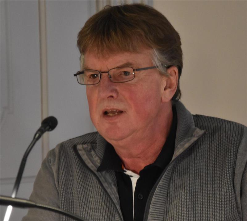 Mann mit Brille und grauem Pullover spricht an einem Mikrofon vor hellem Hintergrund