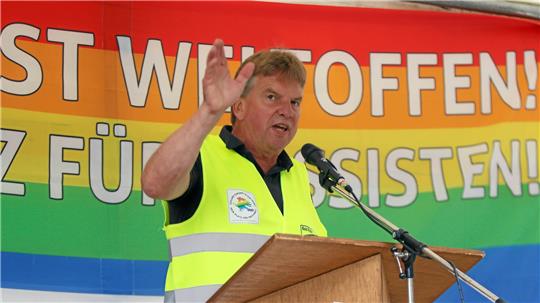 Mann mit gelber Warnweste steht hinter einem Rednerpult und hebt eine Hand vor einem bunten Banner mit großem Text