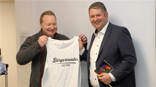 Das Foto zteigt, wie Michael Petrick am Wahlabend ein T-Shirt mit Aufschrift "Bürgermeister" an Martin Schulze erreicht. 