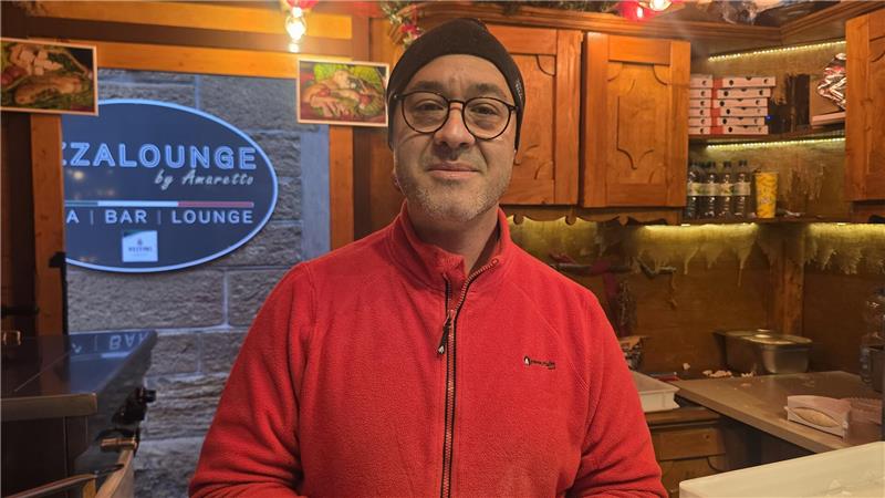 Person in rotem Fleecejacke steht in einer rustikalen Bar mit Holzregalen, Pizzakartons und dekorativen Lichtern.