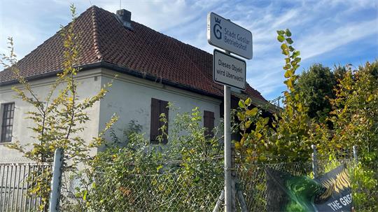 Altes weißes Haus mit rotem Ziegeldach hinter einem Maschendrahtzaun und mit zwei Schildern davor