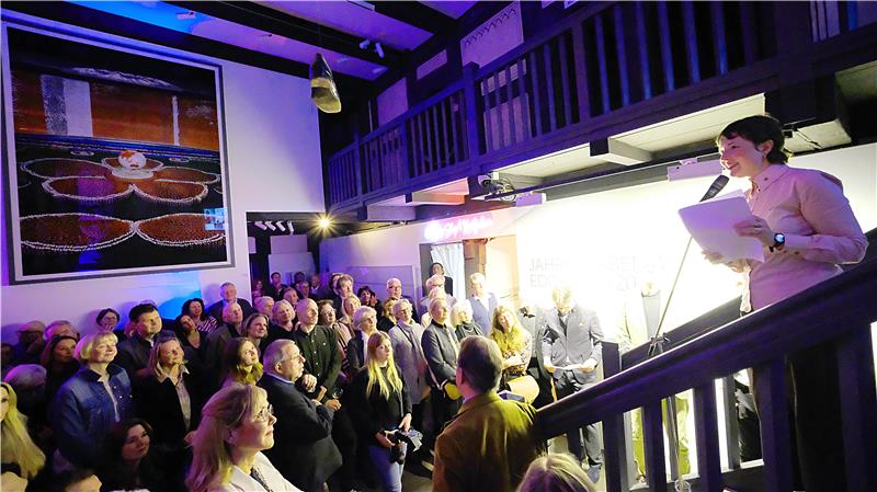 Eine Frau steht auf einer Treppen und spricht in ein Mikrofon. Unter ihr haben sich zahlreiche Menschen versammelt. 