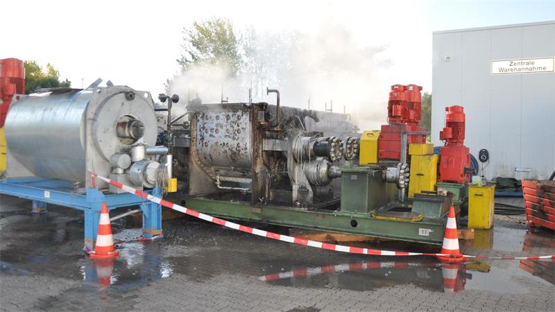 Industriemaschinen mit Dampfaustritt hinter Absperrband und Warnkegeln vor einem Gebäude mit Schild 'Zentrale Warenannahme'