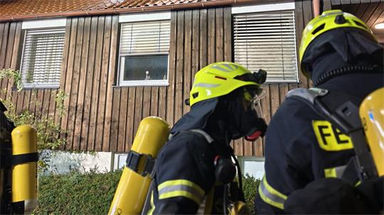 Zwei Feuerwehrleute mit Atemschutzgeräten vor einem zweistöckigen Holzhaus und offenem Fenster im Obergeschoss bei Nacht
