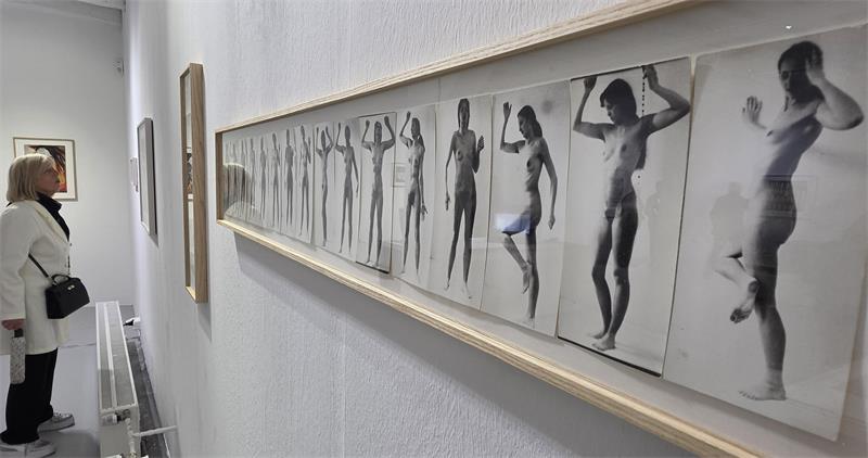 Eine Galerie von schwarz-weißen Nacktaufnahmen hängt an einer weißen Wand. Eine Frau betrachtet die Arbeiten. 