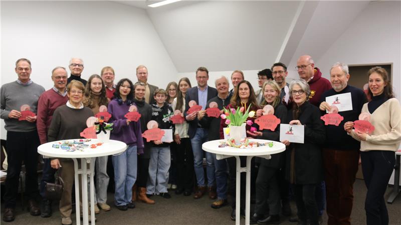 Gruppe von Menschen steht in einem Raum um zwei Stehtische mit Dekoration und hält rosa Papierschweinchen.