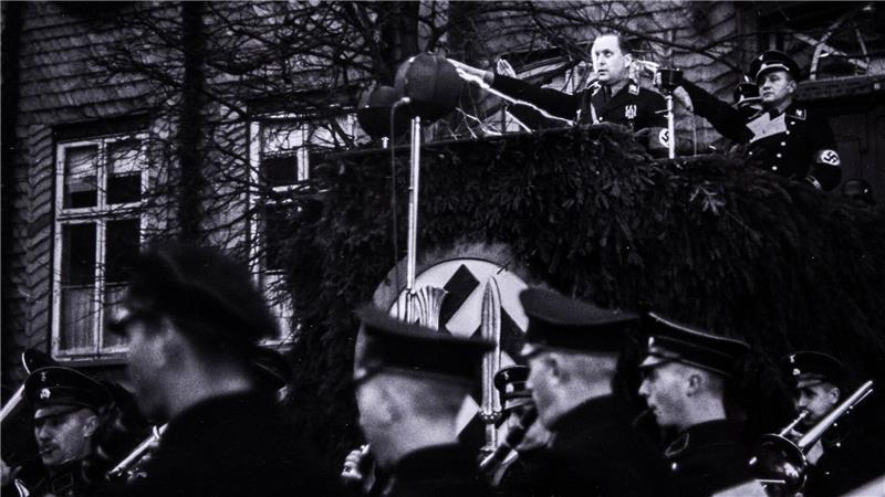Mann in Uniform hält eine Rede von einem mit Tannenzweigen geschmückten Podium vor einer Gruppe uniformierter Männer.