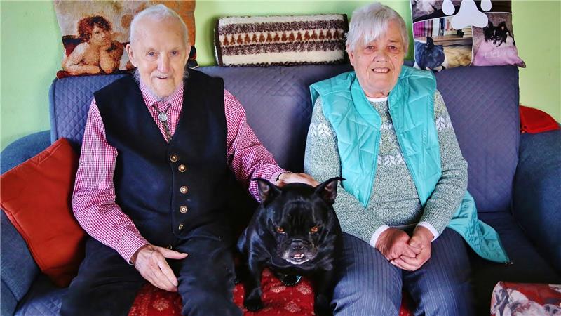 Das Foto zeigt ein Paar zusammen mit ihrem schwarzen Hund auf dem Sofa.