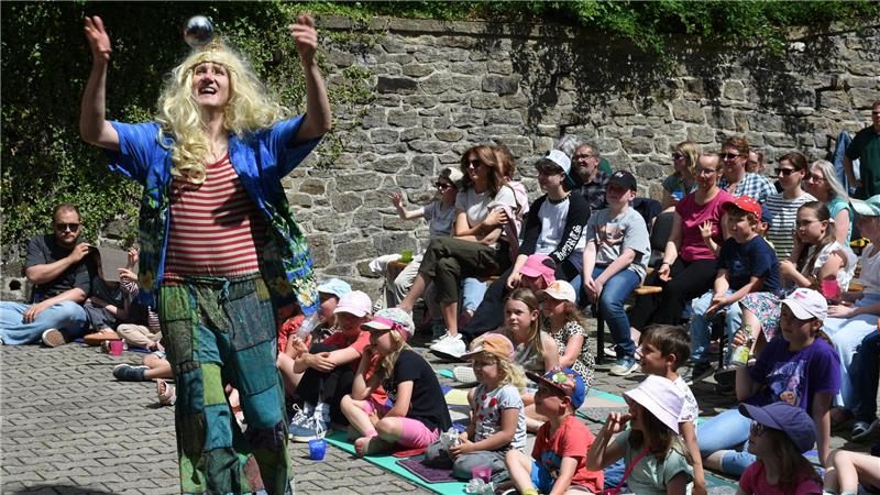 Ein Clown mit blonder Perrücke wirft eine silberne Kugel in die Luft, Kinder sitzen auf dem Boden und auf Bänken und sehen zu. 