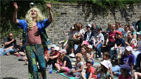 Ein Clown mit blonder Perrücke wirft eine silberne Kugel in die Luft, Kinder sitzen auf dem Boden und auf Bänken und sehen zu. 