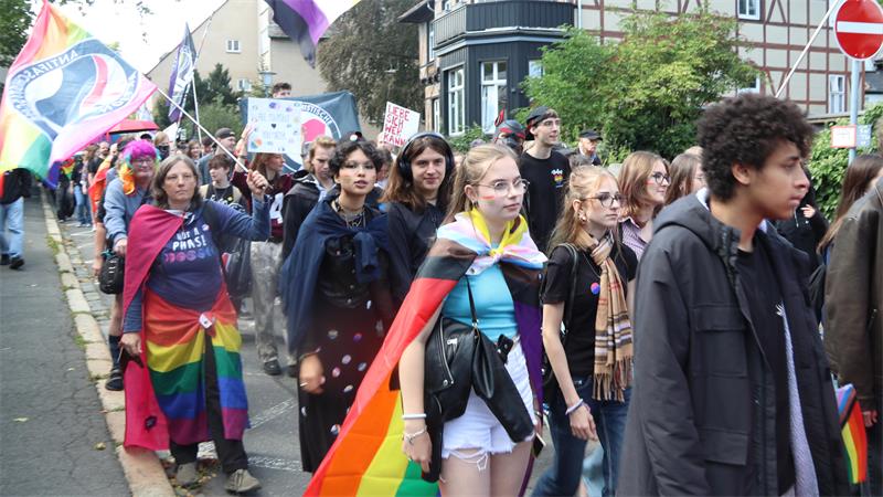 Gruppe von Menschen bei einer Demonstration, einige tragen Regenbogenflaggen und bunte Kleidung, sie gehen eine Straße entlang.