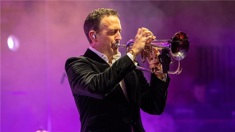 Mann in schwarzem Samtanzug spielt Trompete auf Bühne mit violettem Bühnenlicht.