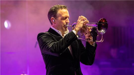 Mann in schwarzem Samtanzug spielt Trompete auf Bühne mit violettem Bühnenlicht.