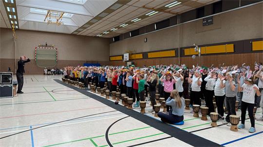 Die Aufnahme zeigt eine große Sporthalle, in der eine größere Gruppe von Kindern gemeinsam mit Trommeln musiziert. Viele der Kinder tragen auffällige Papier- oder Stoffkopfbedeckungen mit Hasenohren. Vor der Gruppe steht eine anleitende Person; im Vordergrund sitzt eine weitere Person auf einem Sitzkissen. Die Halle verfügt über ein Basketballfeld, Tore, Oberlichter und eine umfangreiche Beleuchtungsanlage.