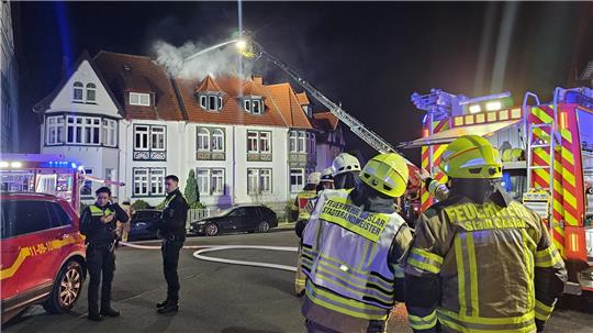 Feuerwehrleute in Schutzkleidung vor einem brennenden Wohnhaus mit Einsatzfahrzeugen und einer ausgefahrenen Drehleiter bei Dunkelheit.