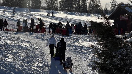 Das Foto zeigt einen vollen Rodelhang und eine Familie im Vordergrund, die ein Kind mit dem Schlitten einen kleineren Hang hinaufziehen.