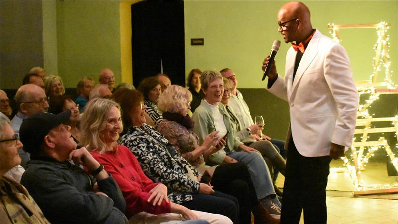 Ein Mann in weißem Sakko steht vor den sitzenden Zuschauern der ersten Reihe und spricht mit ihnen. In der Hand hält er ein Mikrofon. Das Publikum lacht. 